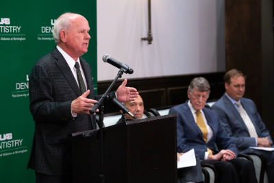 An older man in a suit speaking at a podium in a conference setting with a green UAB Dentistry backdrop with three men in suits sitting in the background